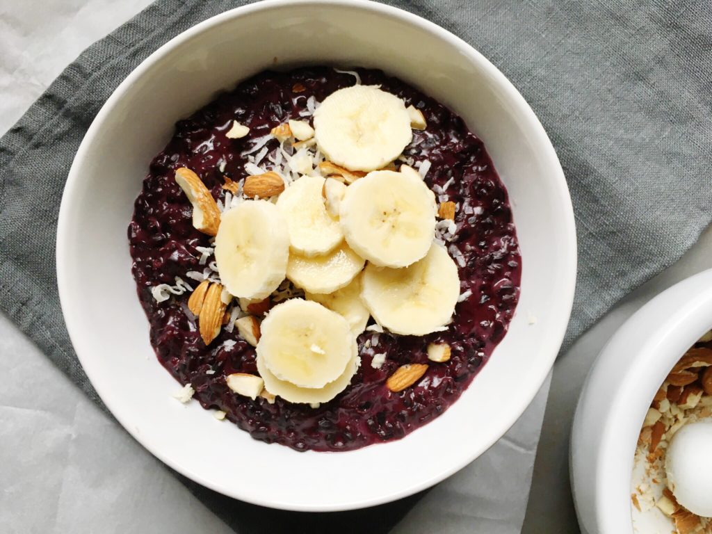 Bowl of coconut black rice breakfast porridge on linen napkin topped with shredded coconut, almonds, and banana