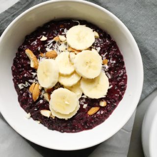 Bowl of coconut black rice breakfast porridge on linen napkin topped with shredded coconut, almonds, and banana