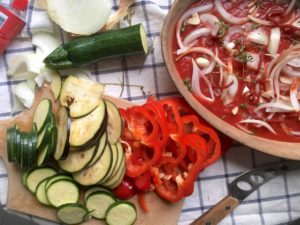 sliced zucchini and red peppers/capsicum on cutting board alongside vintage gratin dish filled with tomato puree and onions