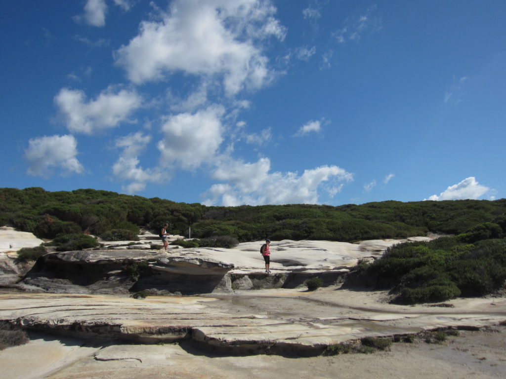 Two people on sandstone and brush cliffs in Royal National Park, with blue sky