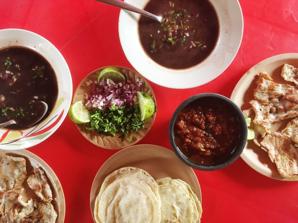 Two bowls of black bean soup, two plates of grilled chicken, a plate of tortillas, salsa, limes, and cilantro on a red table at La Tia de Kaua