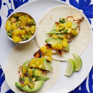 Fish tacos with mango salsa, avocado, and lime, on white plate on blue Otomi background