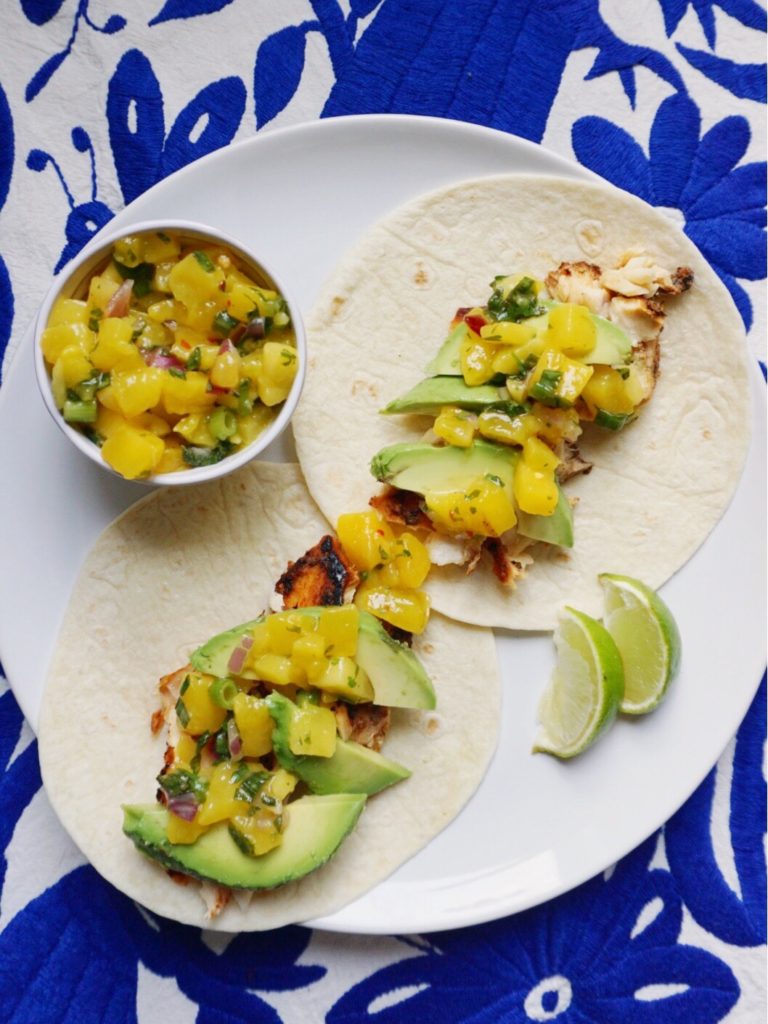 Fish tacos with mango salsa, avocado, and lime, on white plate on blue Otomi background